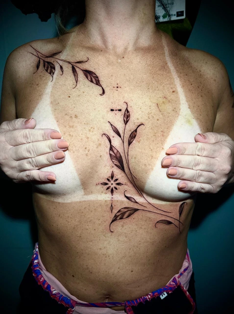 A person from Boca Raton, Florida, with a distinct tan line on their chest, showcases elegant black floral tattoos above and below the line. They are covering their chest with their hands and wearing colorful shorts—true Iron Glory Studio style. in Boca Raton, Florida