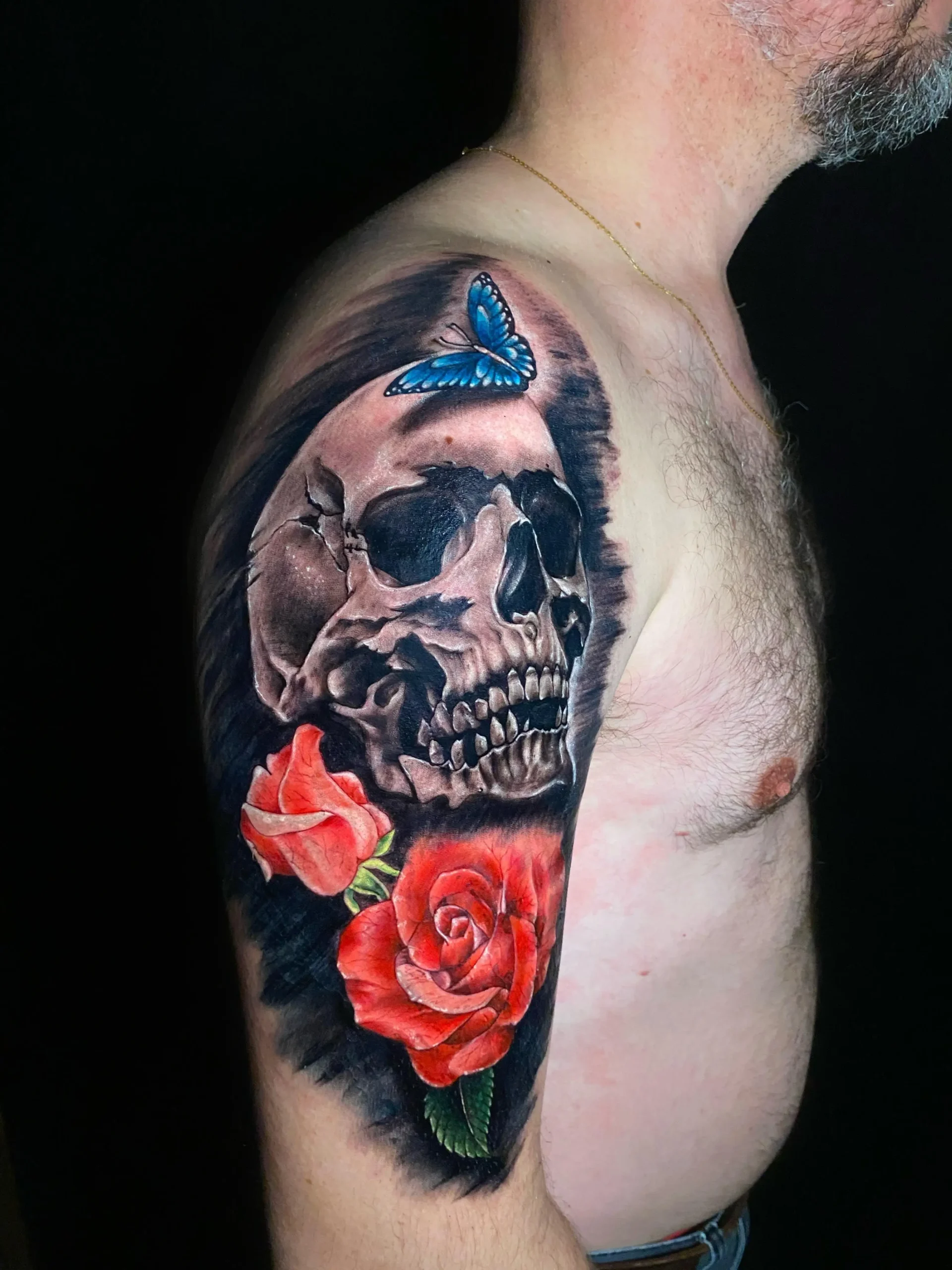 Realistic skull tattoo on the shoulder adorned with red roses and a blue butterfly, high-contrast black and grey ink in Boca Raton, Florida.