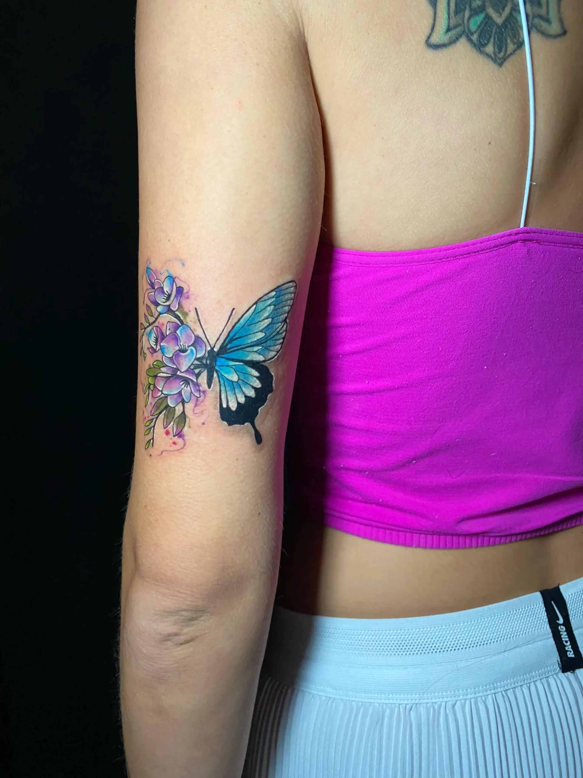 Realistic blue butterfly tattoo with delicate purple flowers on the tricep, feminine and detailed ink by a top Boca Raton artist.