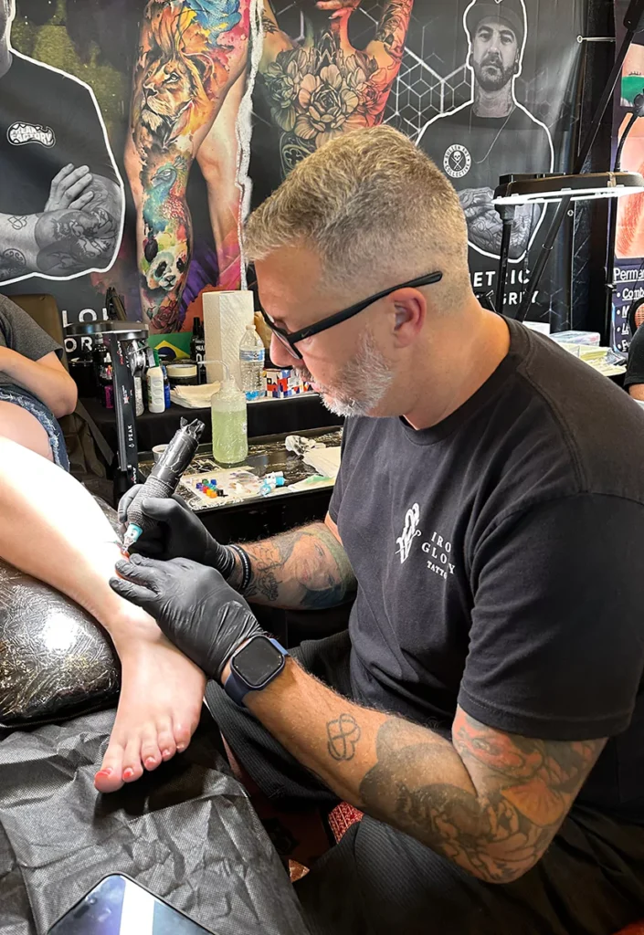 Tattoo artist wearing black gloves and glasses concentrating while tattooing a client's ankle at Iron Glory Tattoo in Boca Raton, Florida. Professional artists offering custom tattoos and specializing in all styles.