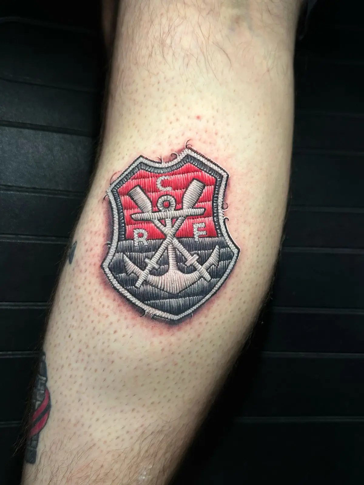 Regatta club shield tattoo featuring an anchor and oars in realistic embroidery patch style, unique textured ink in Boca Raton, Florida.