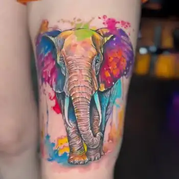 A vibrant, watercolor-style tattoo of an elephant with splashes of bright colors like purple, yellow, pink, and blue on a person’s thigh. The design blends realistic details with artistic, abstract color effects. in Boca Raton, Florida