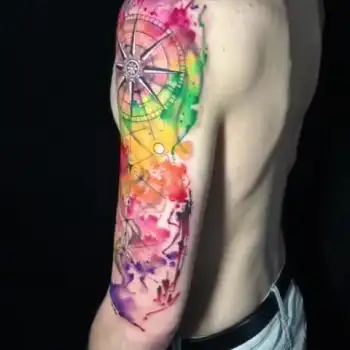 A colorful watercolor-style tattoo of a Ferris wheel with carnival elements covers a persons upper arm, featuring vibrant splashes of pink, orange, yellow, green, and purple. in Boca Raton, Florida