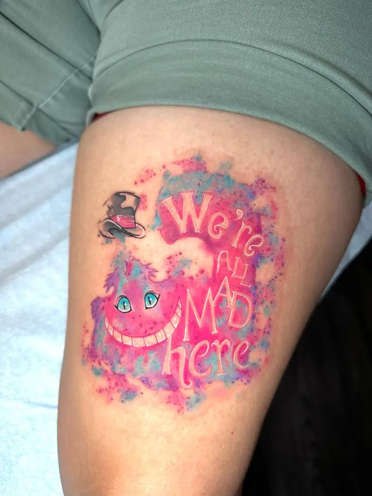 Cheshire Cat watercolor tattoo with 'We're all mad here' quote on the thigh, vibrant Alice in Wonderland ink in Boca Raton, Florida.