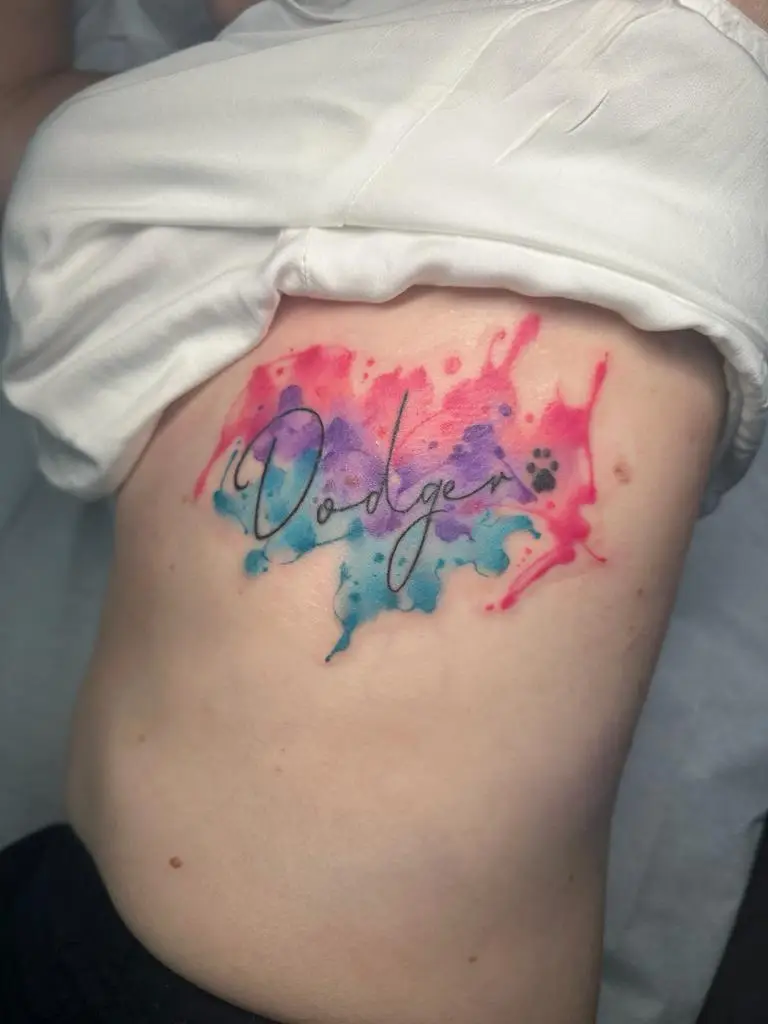 Script name tattoo 'Dodger' with a paw print and blue/pink watercolor splash on the ribs, pet memorial ink in Boca Raton, Florida.