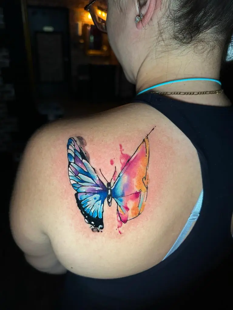 Artistic butterfly tattoo on the shoulder blade, split between realistic blue wings and abstract orange watercolor, inked in Boca Raton, Florida.