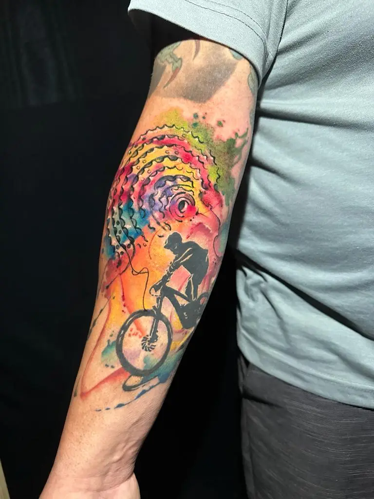 Mountain biker silhouette tattoo with colorful watercolor gears and wheels on the forearm, artistic sports ink in Boca Raton, Florida.